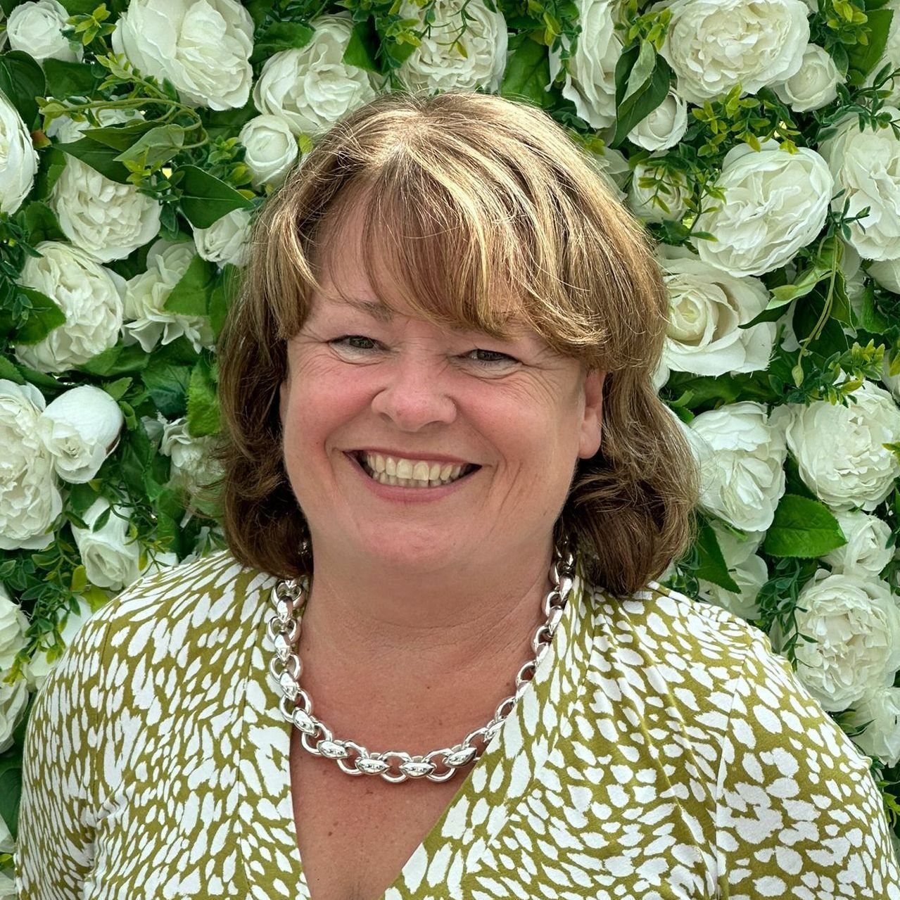 A woman wearing glasses and a necklace is smiling in front of a wall of flowers.