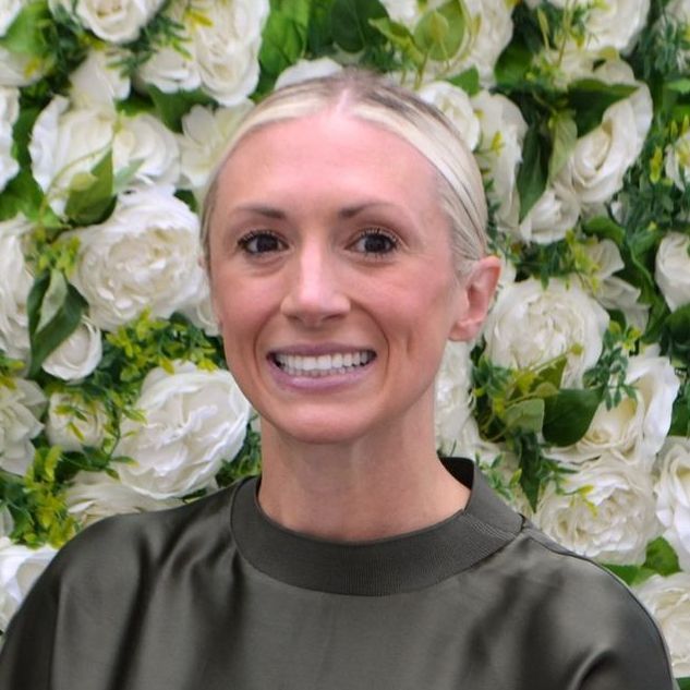 An elderly woman wearing glasses and a necklace smiles in front of a wall of white roses