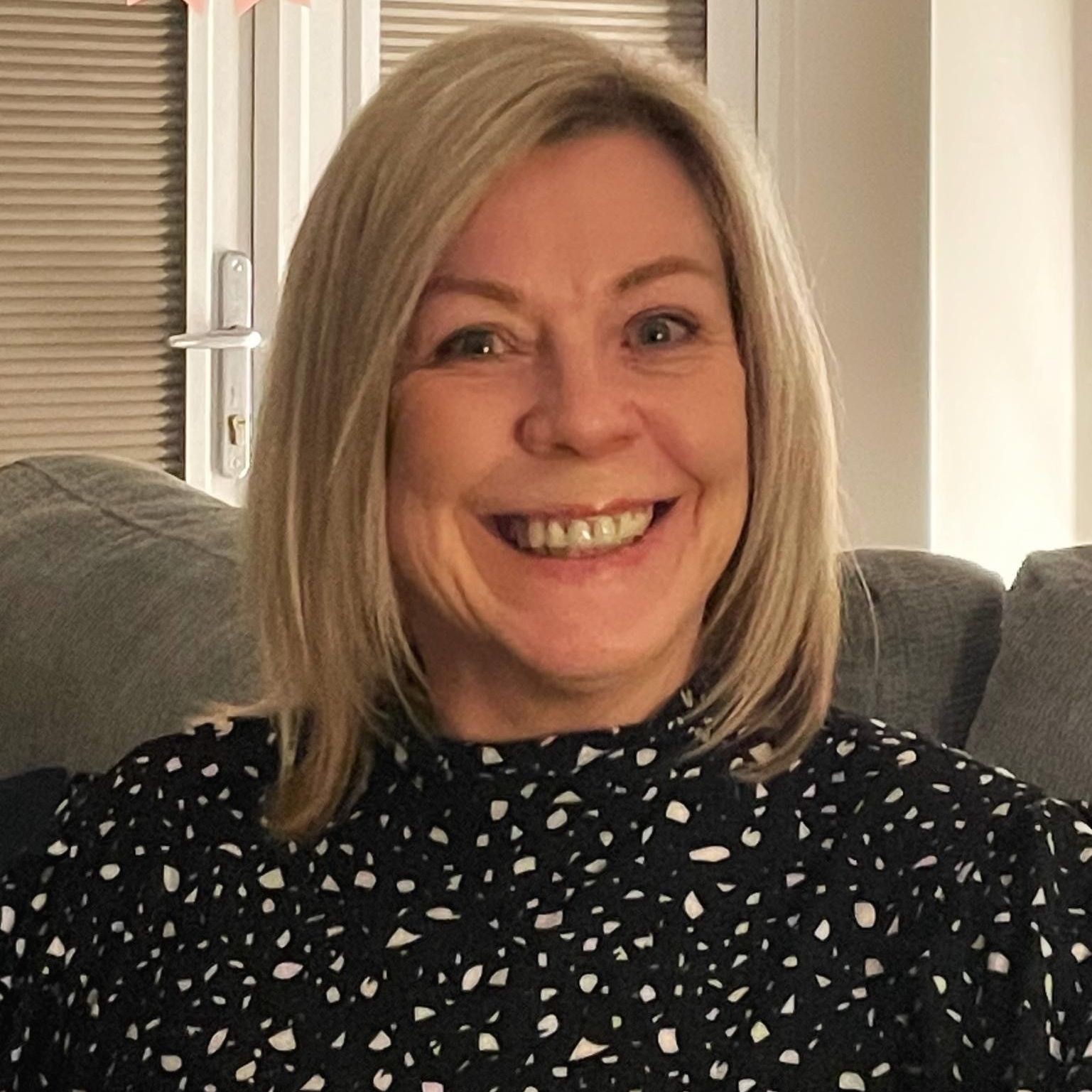 A woman in a yellow jacket is smiling in front of a wall of white flowers