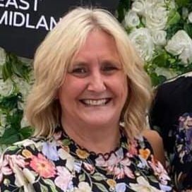 A woman in a yellow jacket is smiling in front of a wall of white flowers