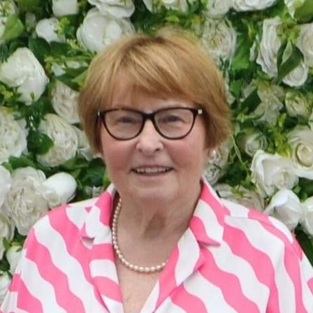 A woman in a yellow jacket is smiling in front of a wall of white flowers