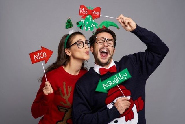 Couple in festive sweaters holding holiday signs and mistletoe props, smiling against a gray background