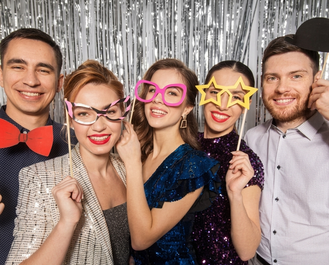 Five people posing with colorful novelty glasses in front of a silver tinsel backdrop