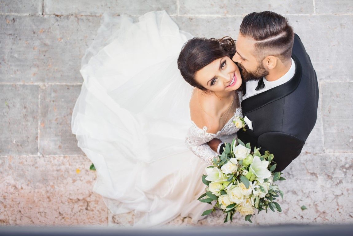 Bride in white dress and groom in black suit embracing and kissing from above, holding a bouquet.