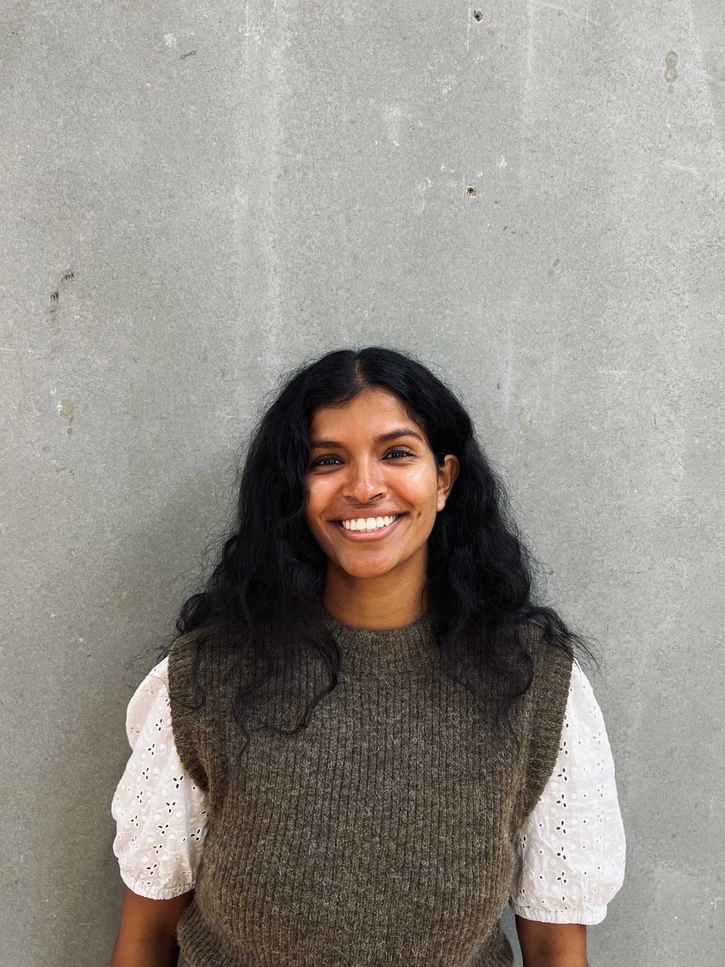 En kvinde i brun vest og hvid skjorte smiler foran en grå væg.