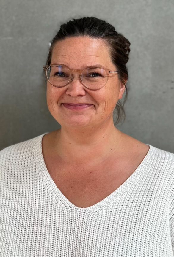 En kvinde iført briller og en blå skjorte smiler foran en grå væg.