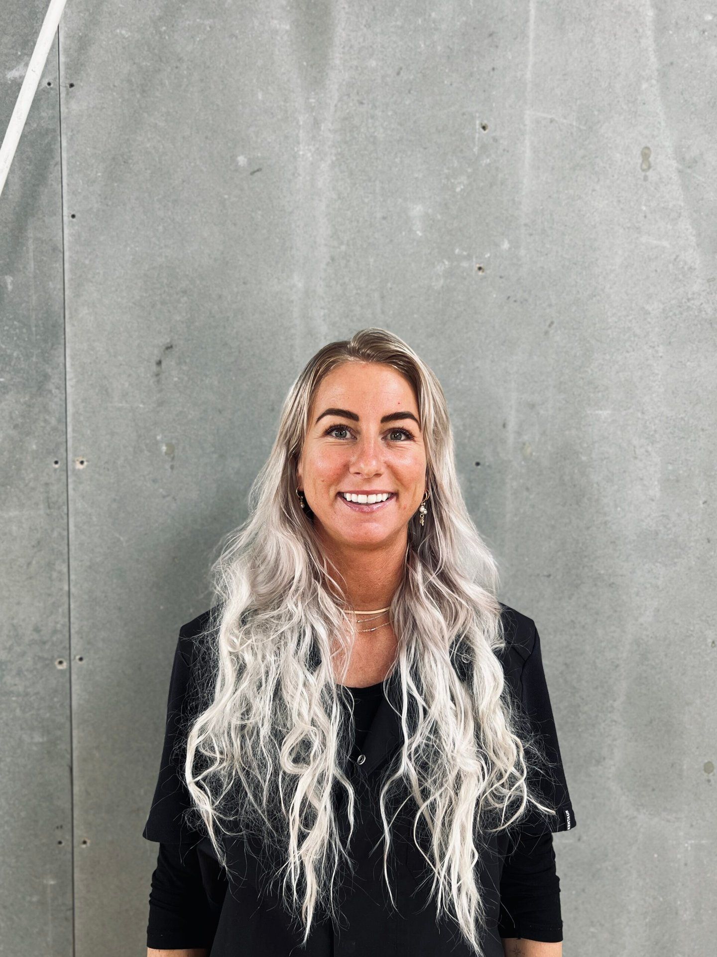 En kvinde med langt blondt hår smiler foran en grå væg.