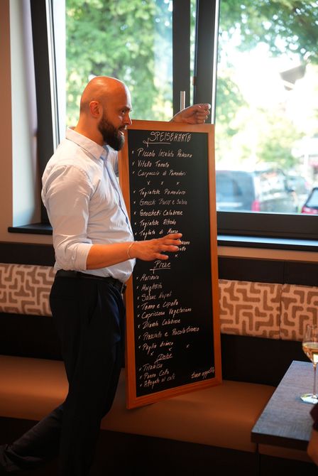 Ein Mann präsentiert sich neben einer hohen, schwarzen Speisekarte in einem hellen Restaurant an großen Fenstern