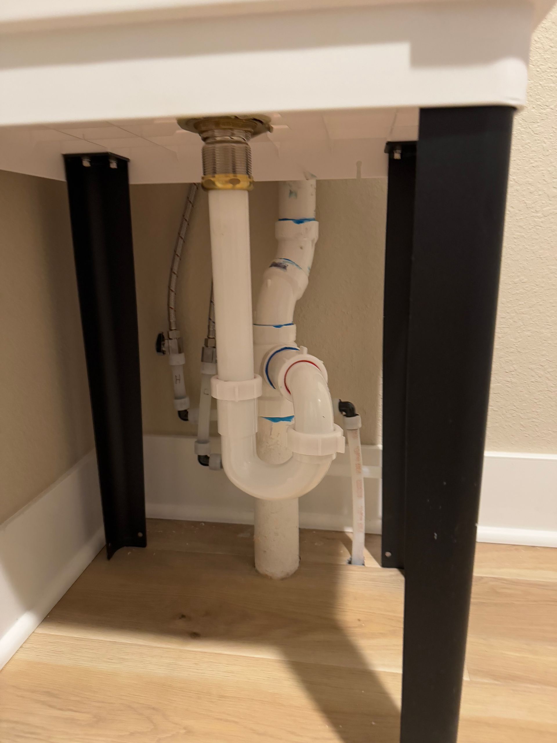 Underside of a utility sink with white plumbing and black supports; against a light-colored wall and floor.