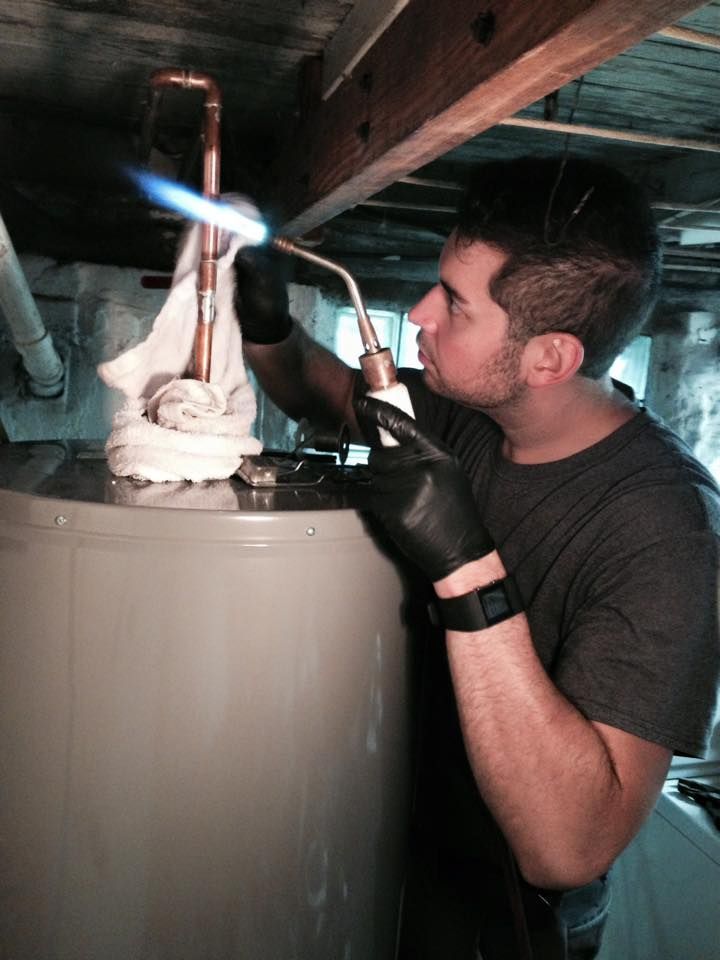 Person soldering copper pipe on top of a water heater with a torch; inside, dimly lit.