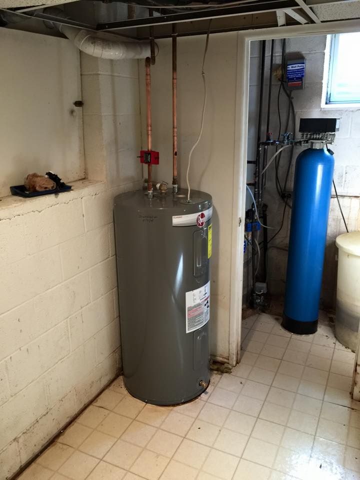 Water heater and water softener in a basement setting. Gray tank with copper pipes, blue softener, white tile floor.