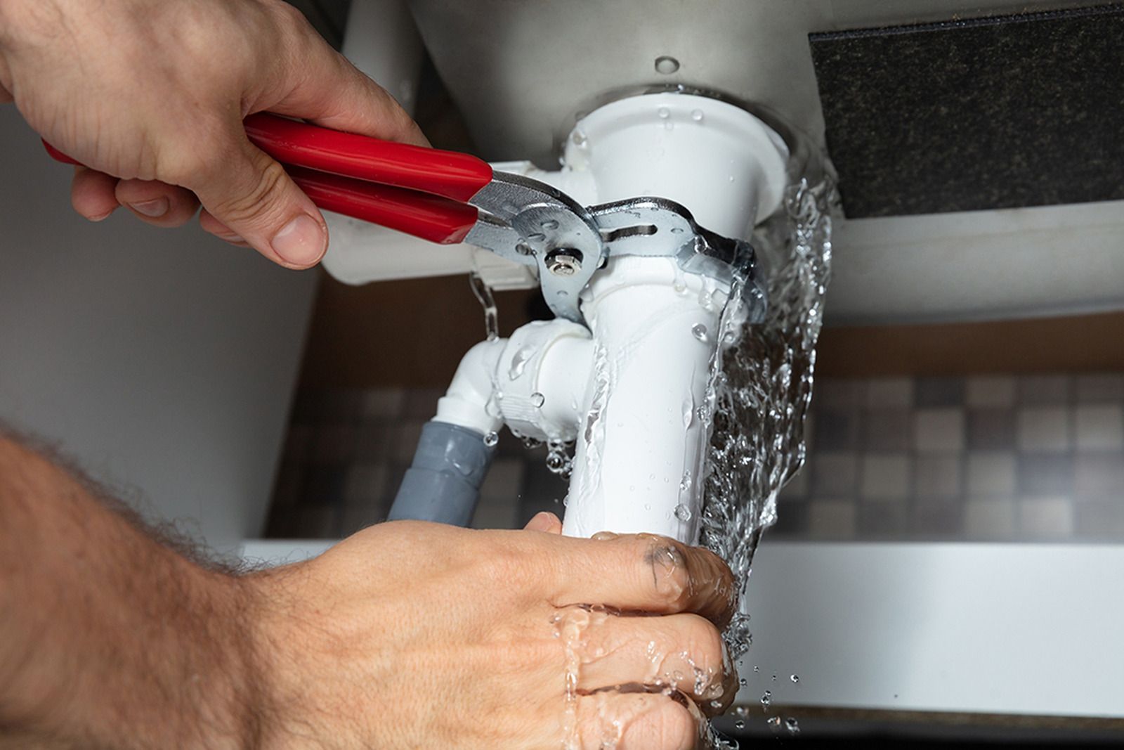 Plumber uses pliers to tighten a white pipe under a sink; water leaks out.