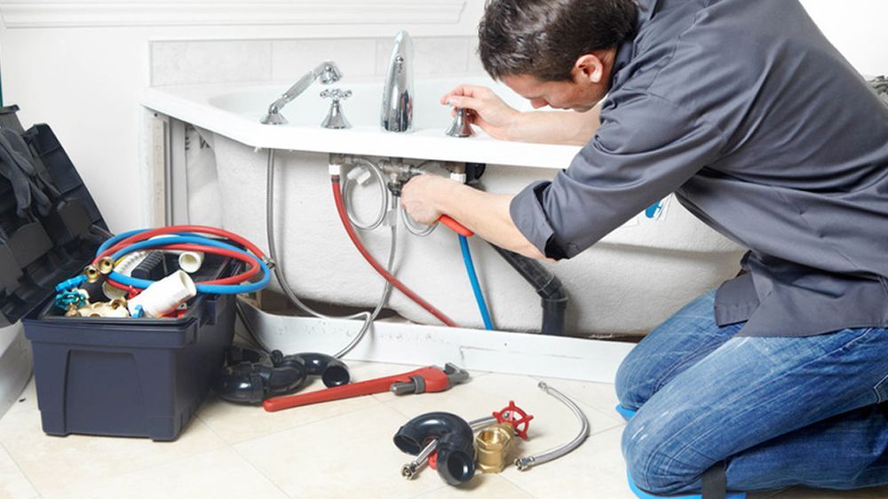 Plumber kneeling, working on pipes under a bathtub. Toolbox with tools beside him.