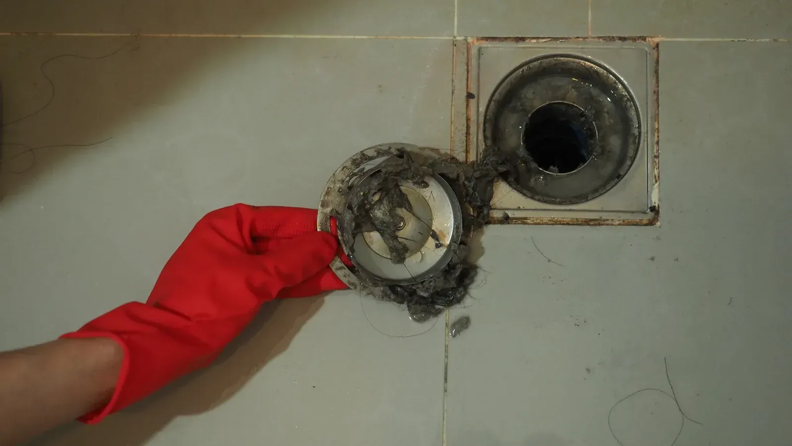 Hand in a red glove holding a clogged drain cover over a shower drain. Gray tile background.