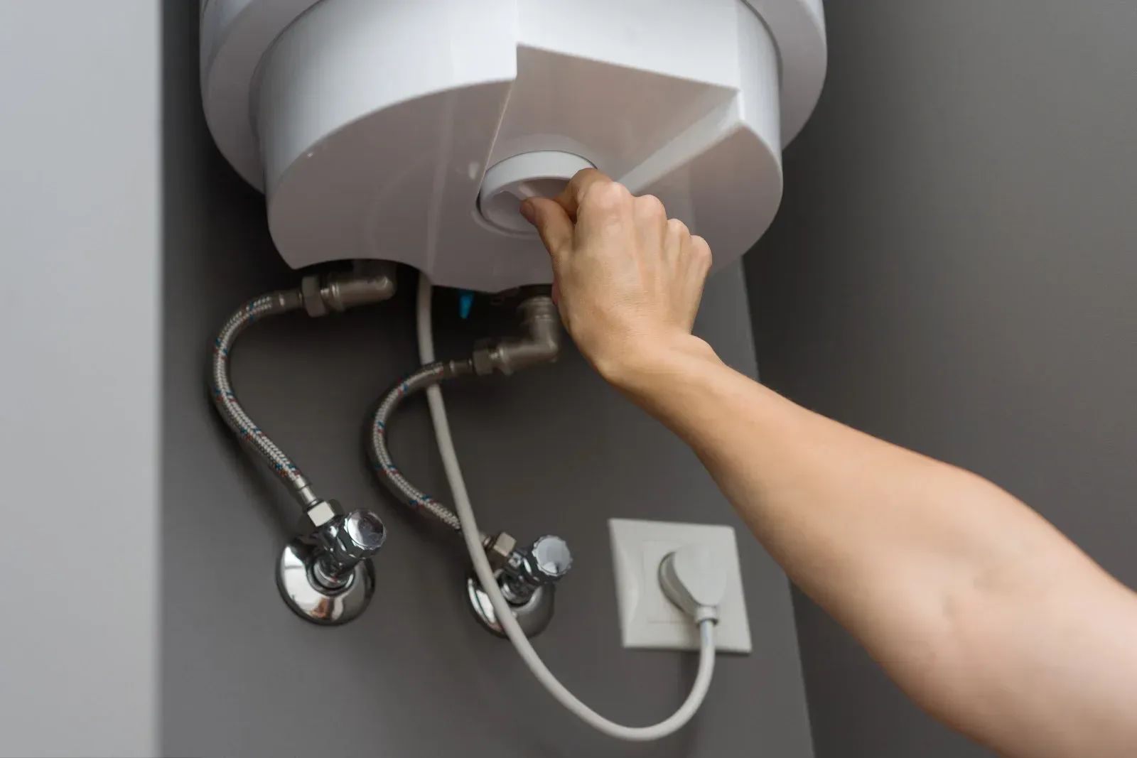 A person's hand turning a knob on a white water heater, connected to pipes on a gray wall.
