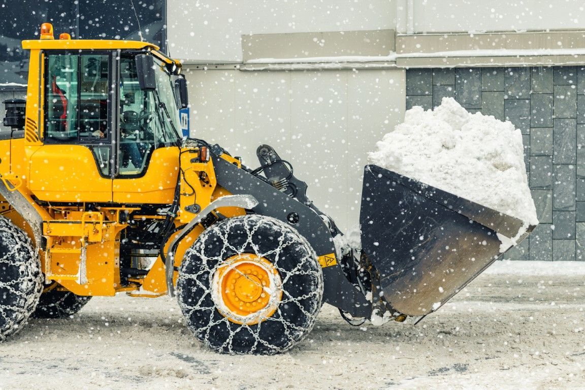 Yellow front loader with chained tires moving a large pile of snow during heavy snowfall in an urban area.