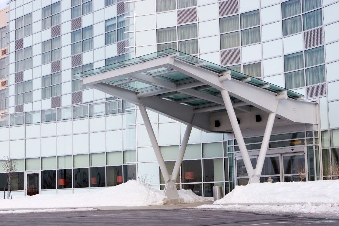 Modern building entrance with glass canopy and snow piled on the ground, featuring large windows and sliding doors.