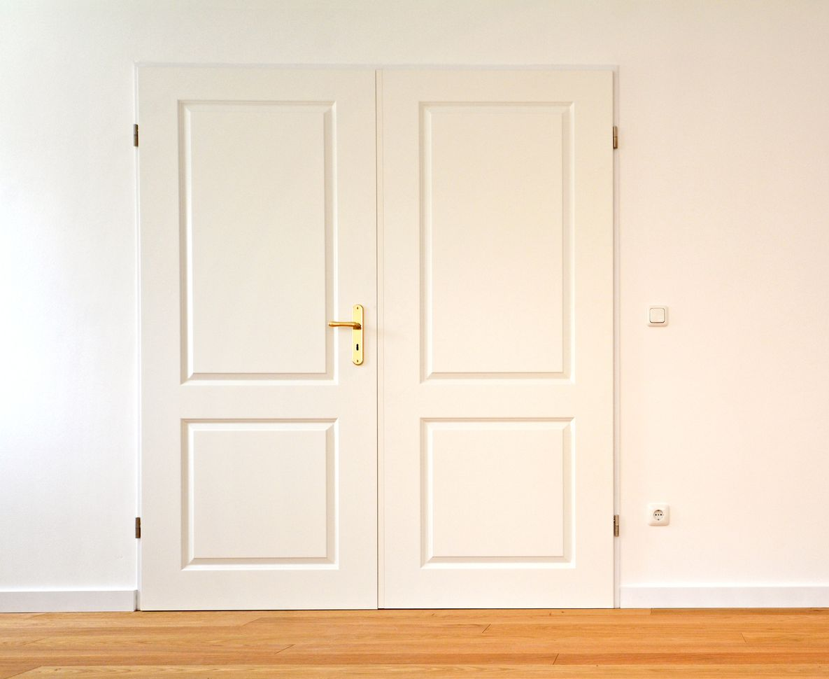 Weiße Doppeltüren mit goldenen Griffen sind in eine schlichte, helle Wand über einem Holzboden eingelassen.