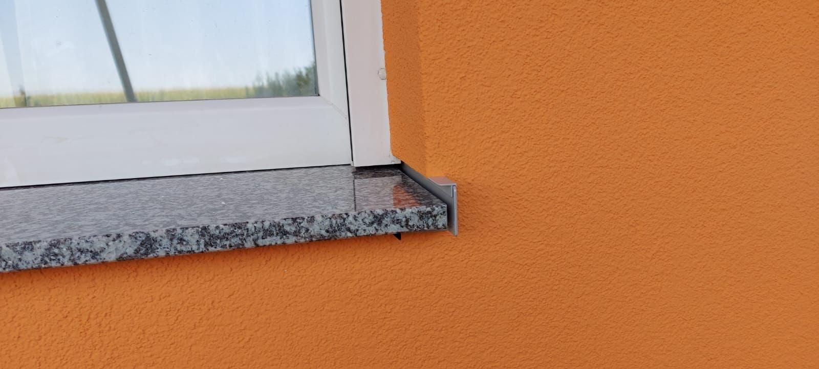 Eine Fensterbank aus Granit, an orangefarbenen, strukturierten Außenwand mit einer Metallabschlusskappe angebracht ist.
