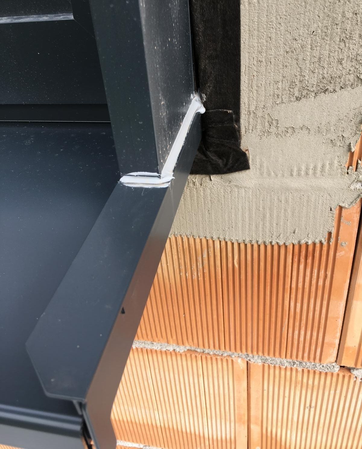 Nahaufnahme einer dunklen Metallfensterbankfuge, die mit weißem Dichtmittel an unfertigen Ziegel- und Putzwand 