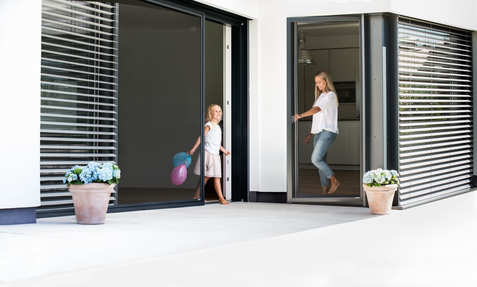 Kind mit Luftballons in der Hand verlässt eine Schiebetür zur Terrasse, während eine Person in angrenzenden Türrahmen steht.