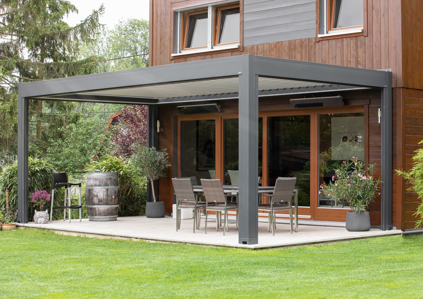 Eine moderne, dunkelgraue Metallpergola über einer Steinterrasse mit Essmöbeln im Freien vor einer Holzhausfassade.