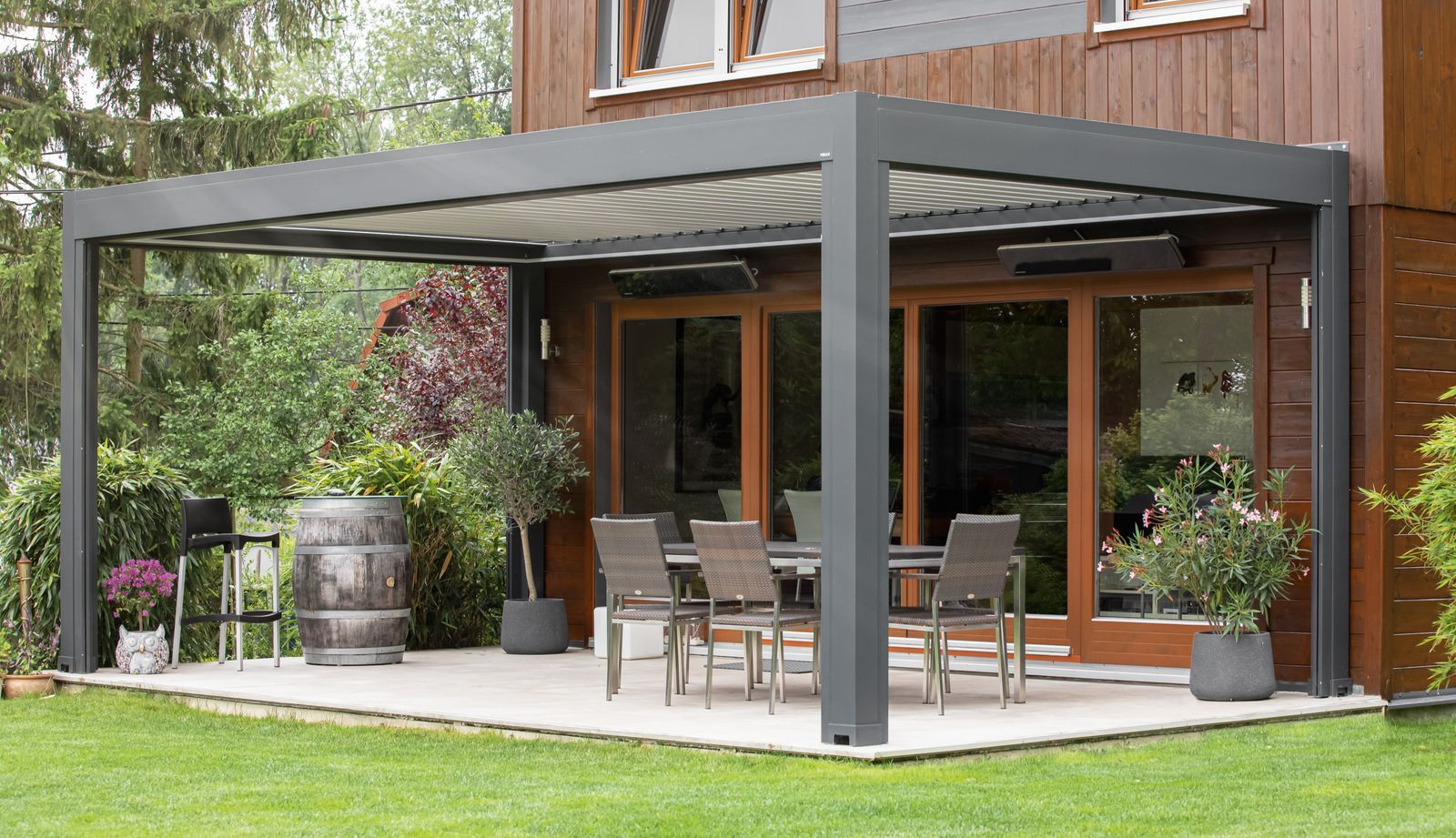 Vor einem Holzhaus steht auf einer Steinterrasse eine dunkle Metallpergola, auf der Gartenmöbel und Topfpflanzen stehen.
