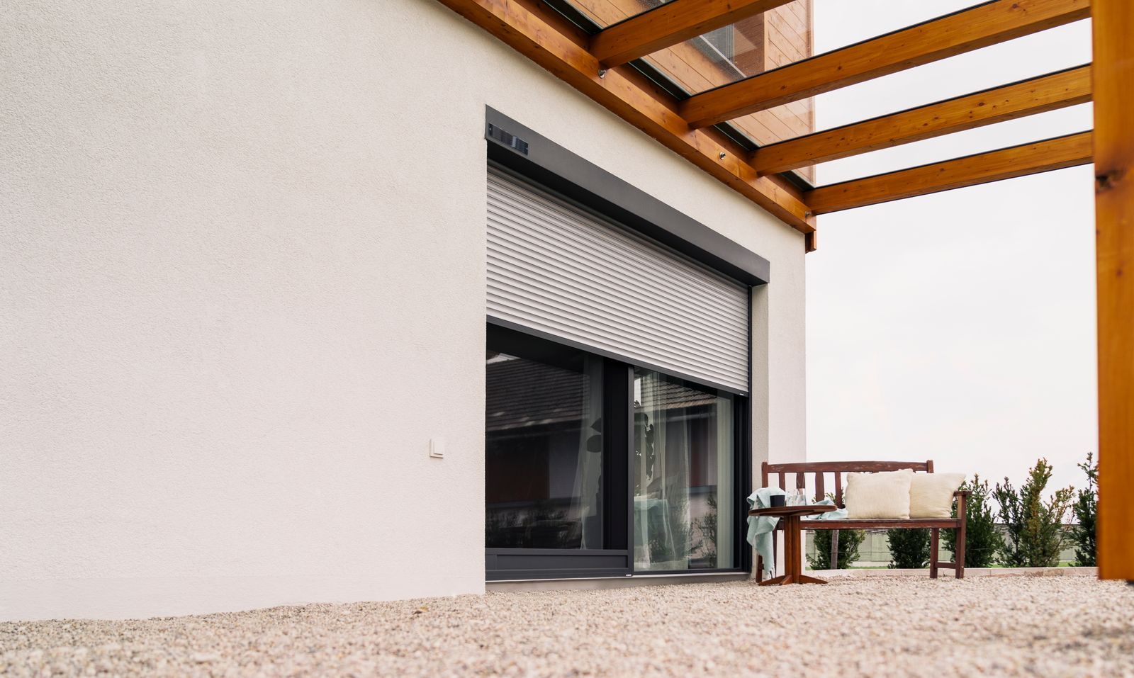 Eine moderne Hausfassade mit heller Stuckwand, einem teilweise geschlossenen Rollladen und einer hölzernen Terrassenpergola.