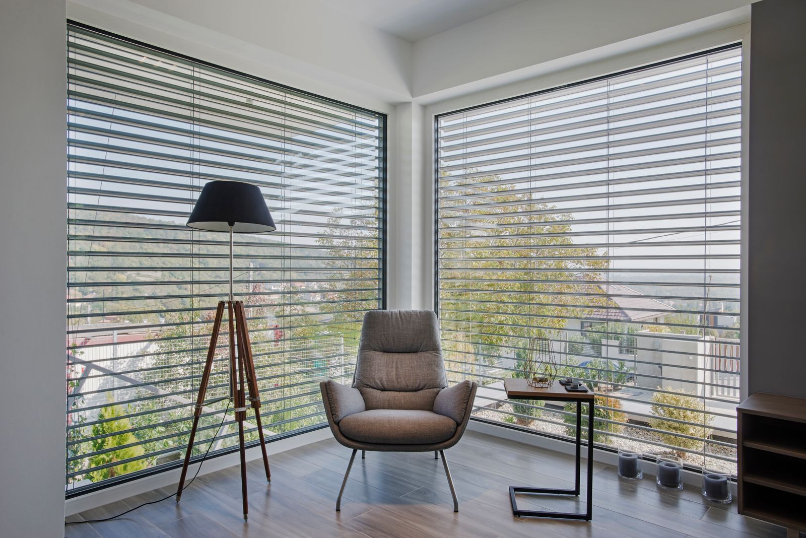 gemütliche Ecke mit einem grauen Sessel, einer Stehlampe mit Dreibeinständer , vor bodentiefen Fenstern mit Jalousien.