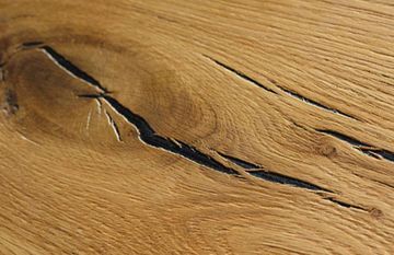 Nahaufnahme der hellbraunen Holzmaserung mit einem dunklen, natürlichen Astloch, durch das tiefe, verfüllte Risse verlaufen.