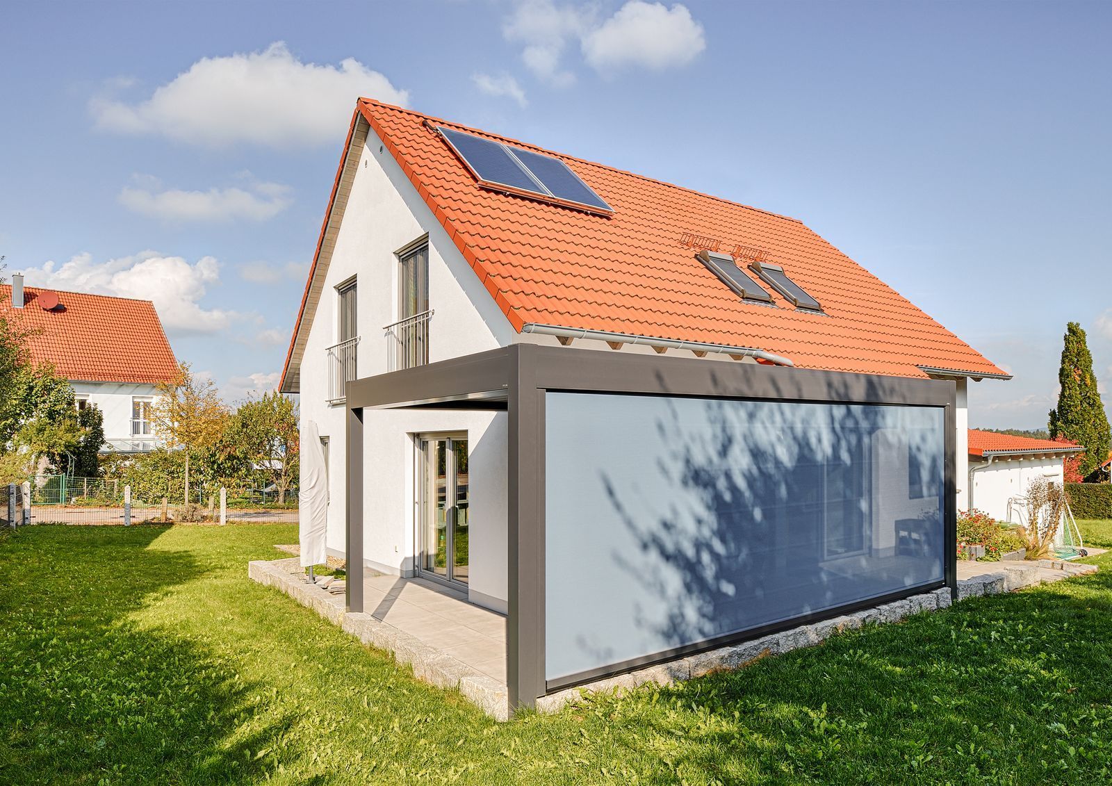 weißes zweistöckiges Haus mit rotem Ziegeldach und Solarpaneelen, mit  überdachten Terrasse mit blauer Seitenwand.