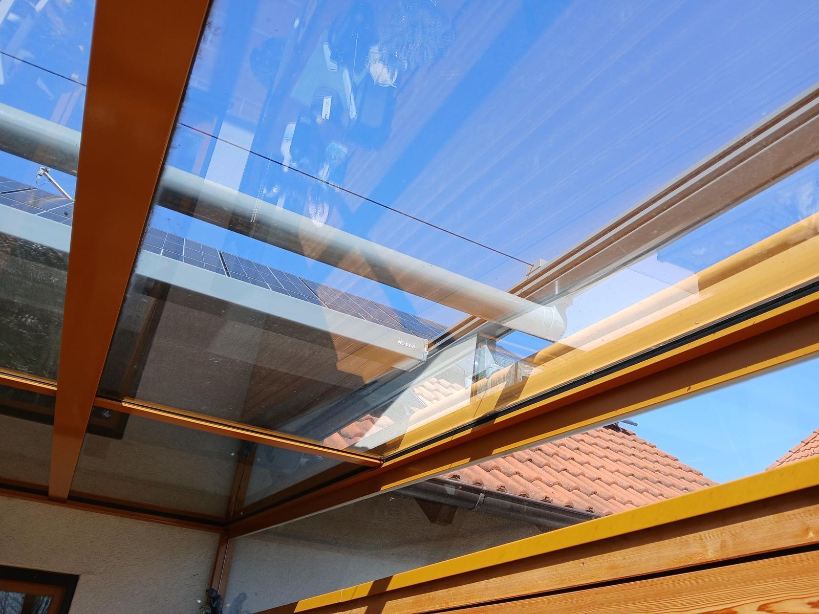 Blick durch das Glasdach eines Wintergartens mit Holzrahmen, draußen sieht man ein Ziegeldach und den blauen Himmel.