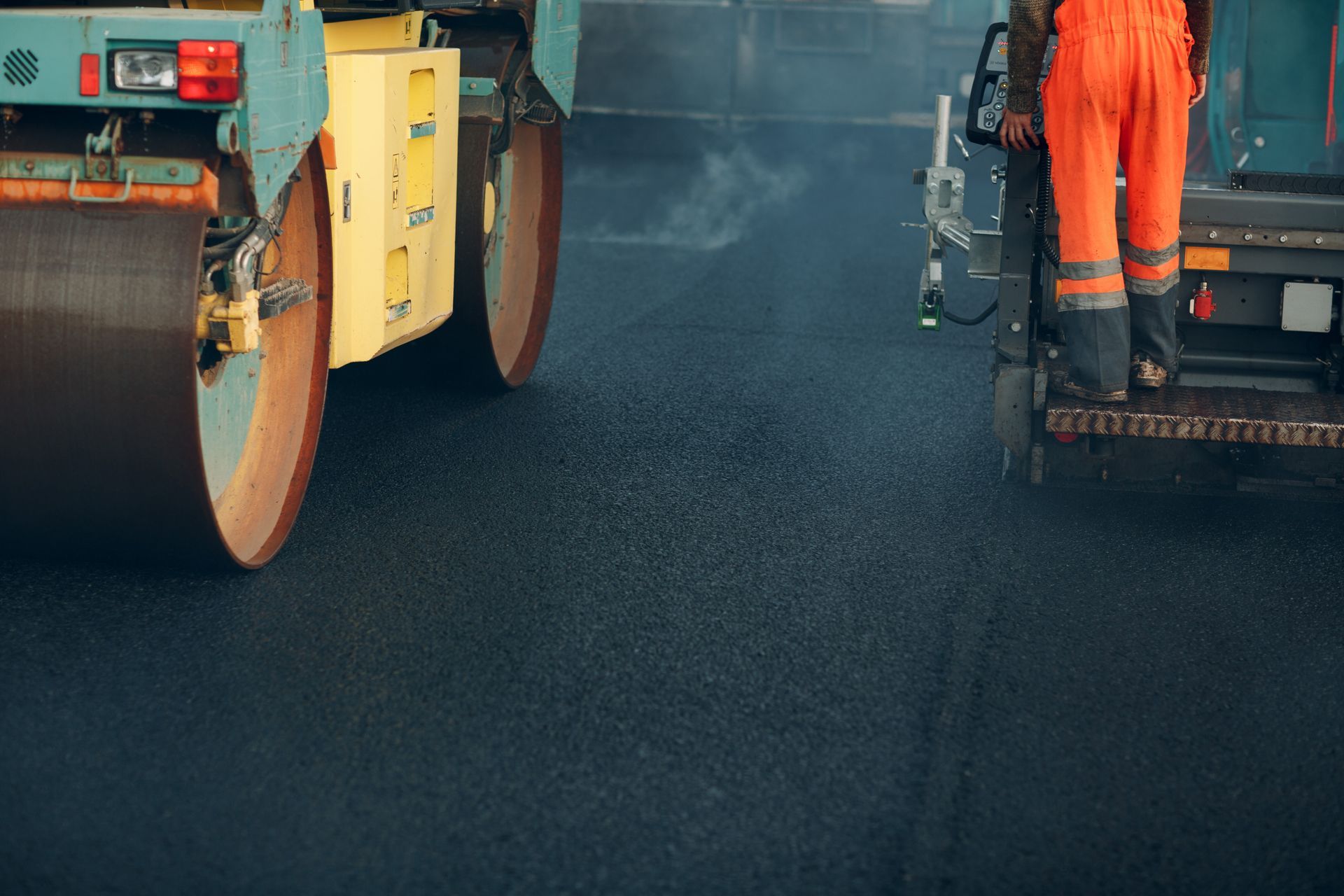 Fresh asphalt compacted by roller, worker in orange gear paving a smooth new road.