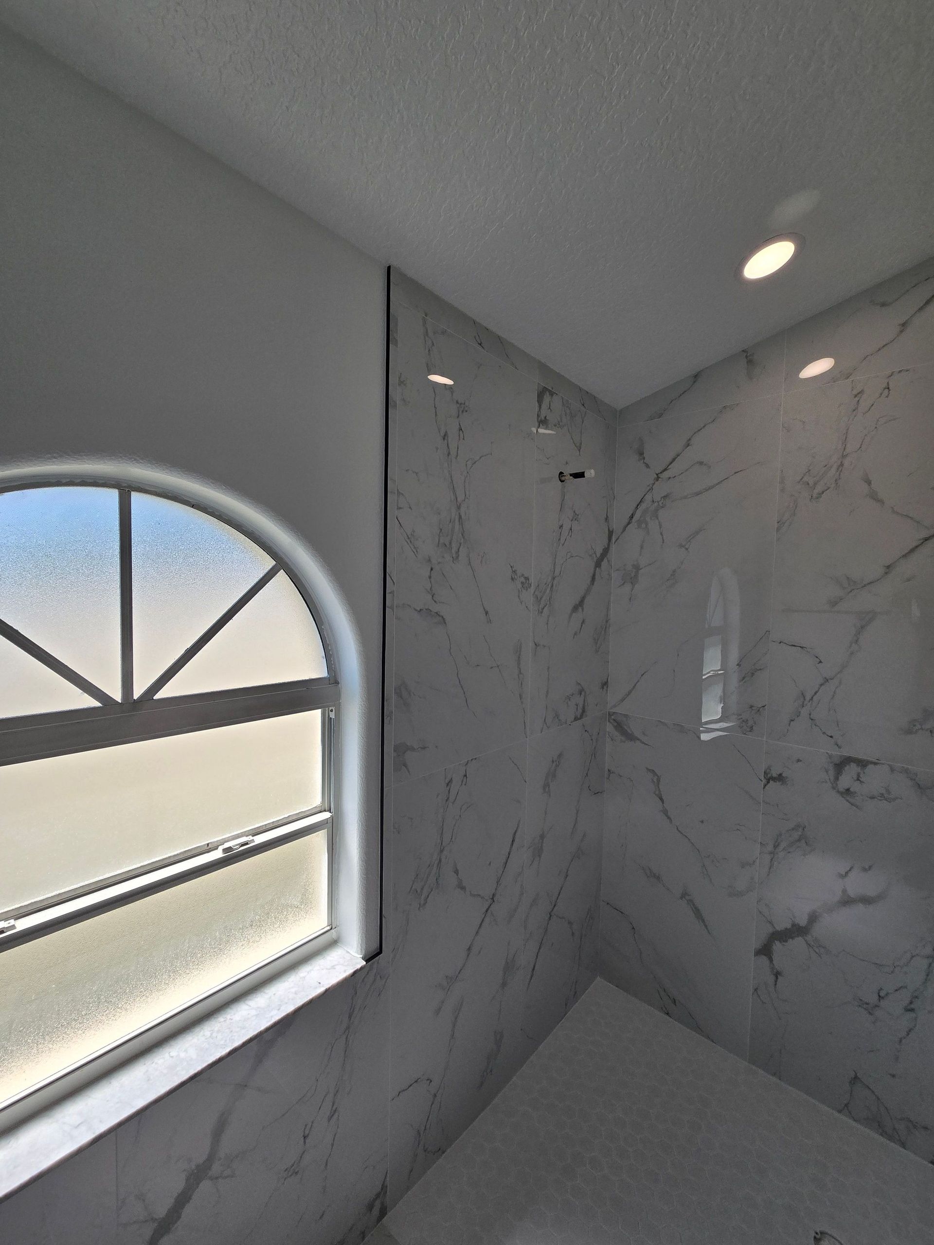 Small marble-tiled shower corner with an arched window and recessed light.