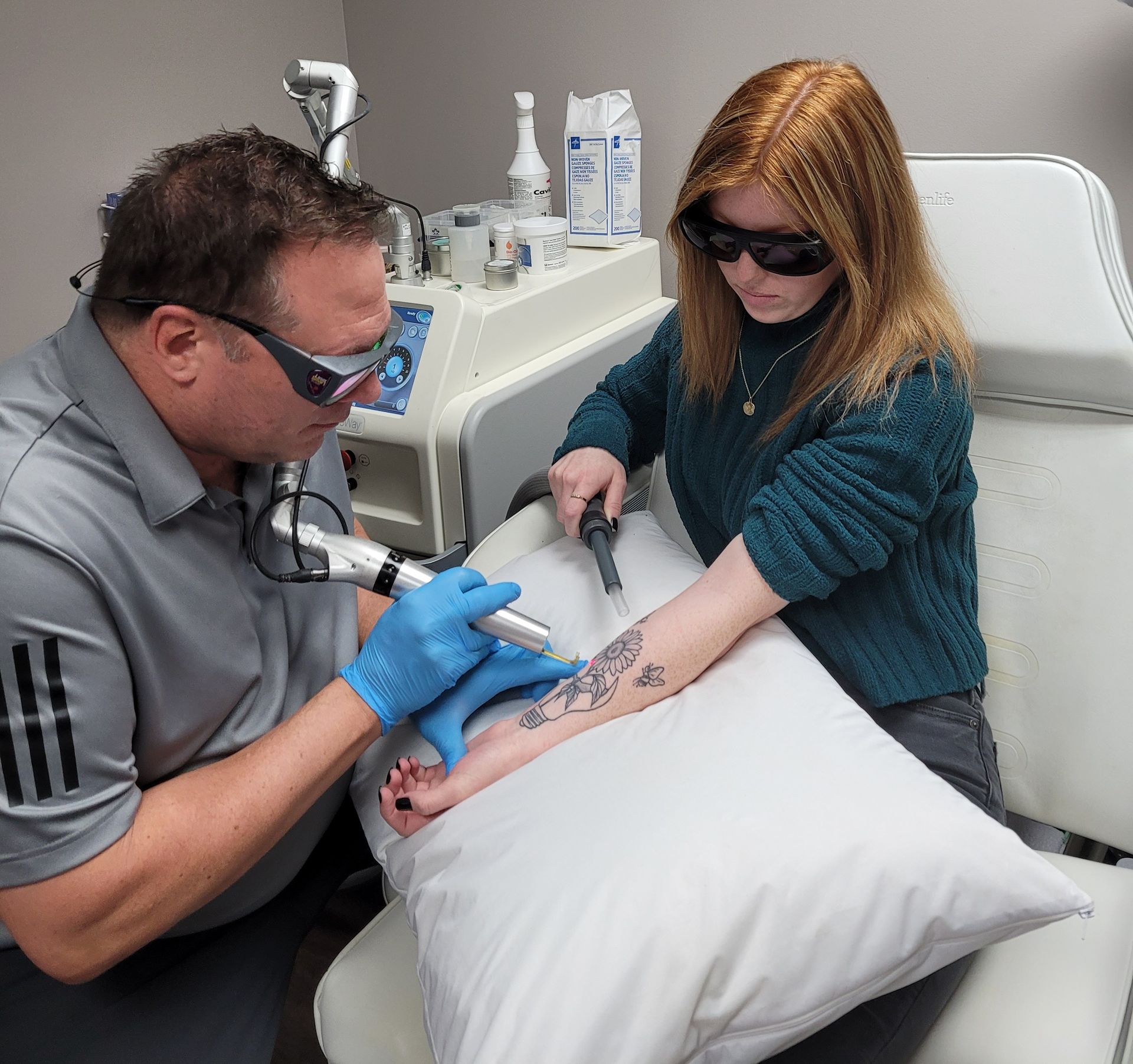 Man using laser on a woman's tattoo on her arm; both wearing protective eyewear in a clinic.