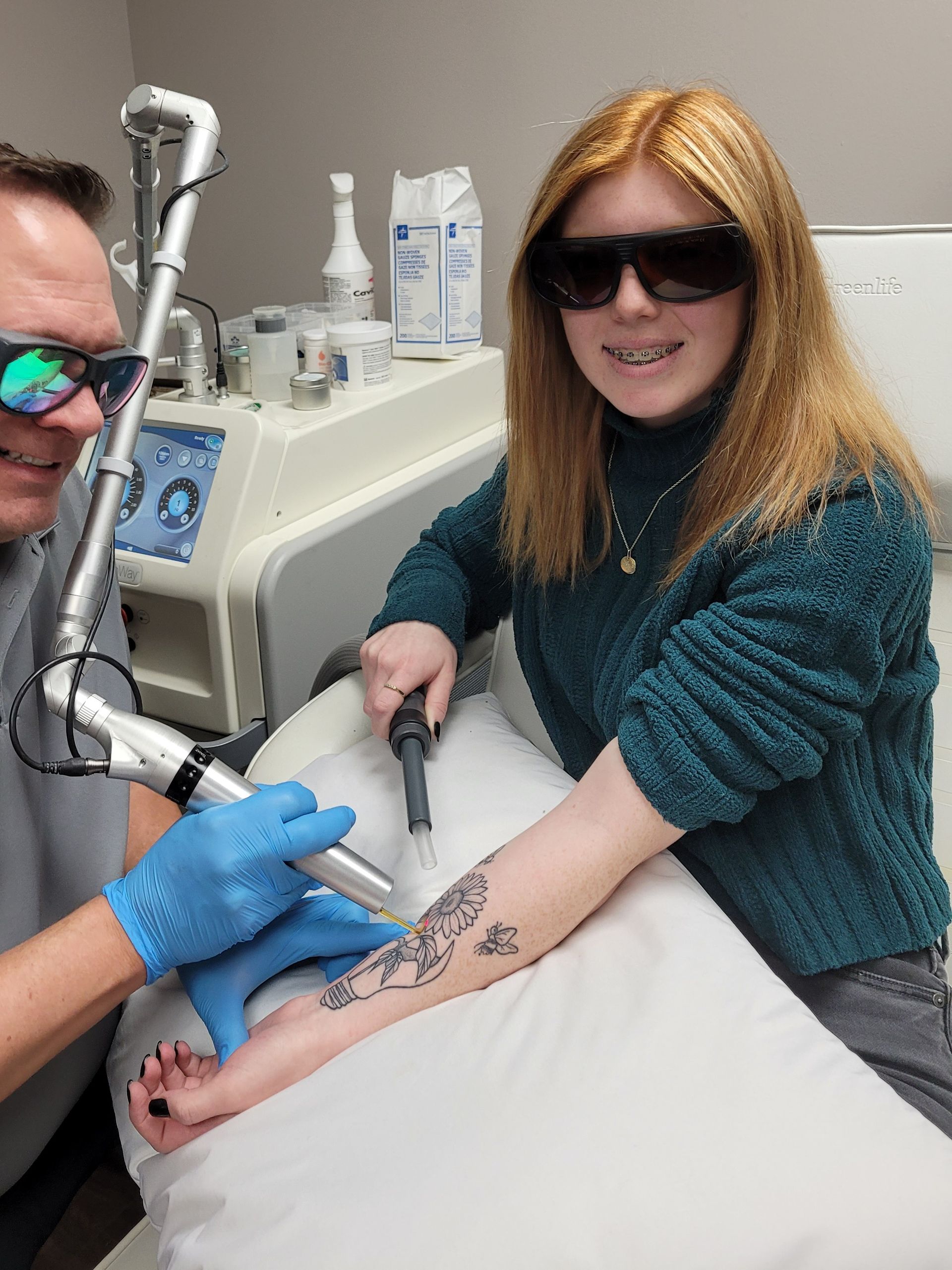Tattoo removal treatment on a seated person wearing sunglasses in a clinic, with a laser device applied to the forearm.