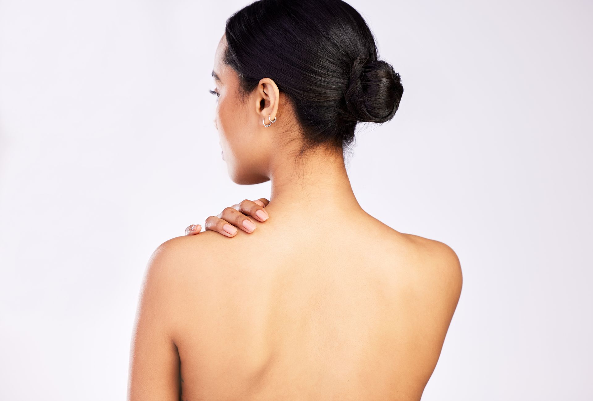 Woman's bare back, touching her shoulder. Dark hair in a bun. Against a plain white backdrop.