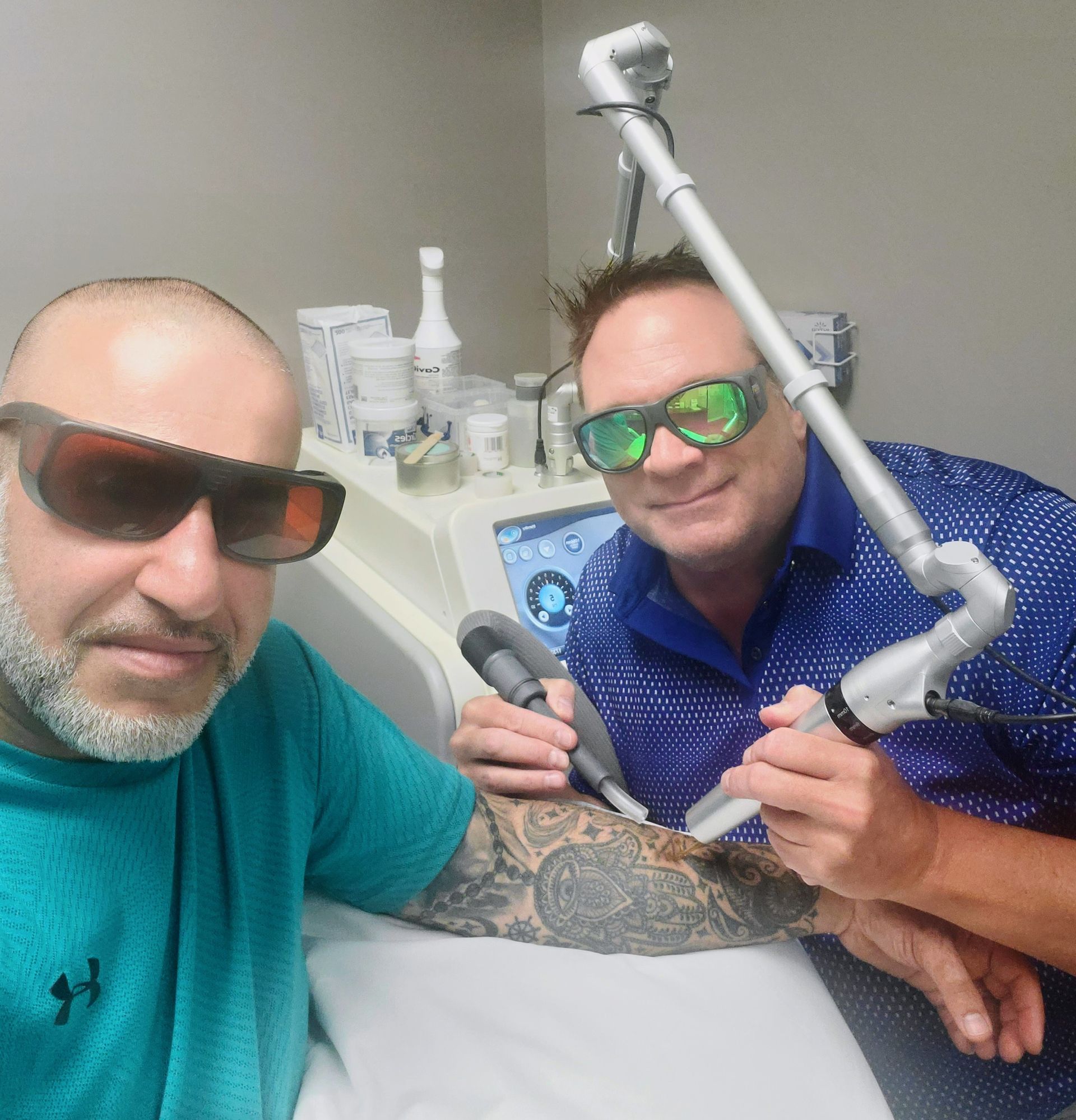Two men in protective eyewear; one receiving tattoo removal on arm in a medical setting.