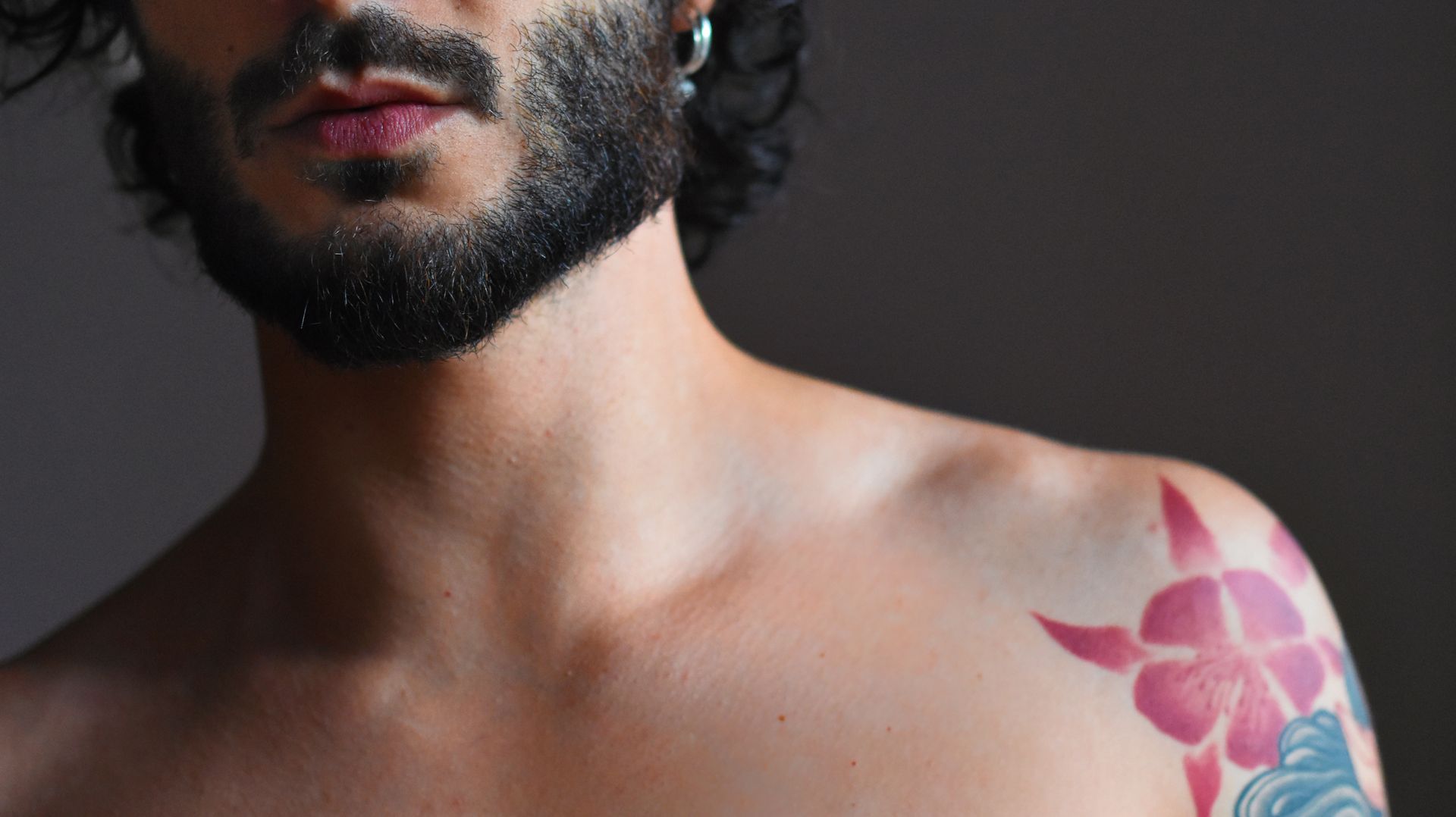Man's shoulder and face with a pink flower tattoo. Dark beard and skin.