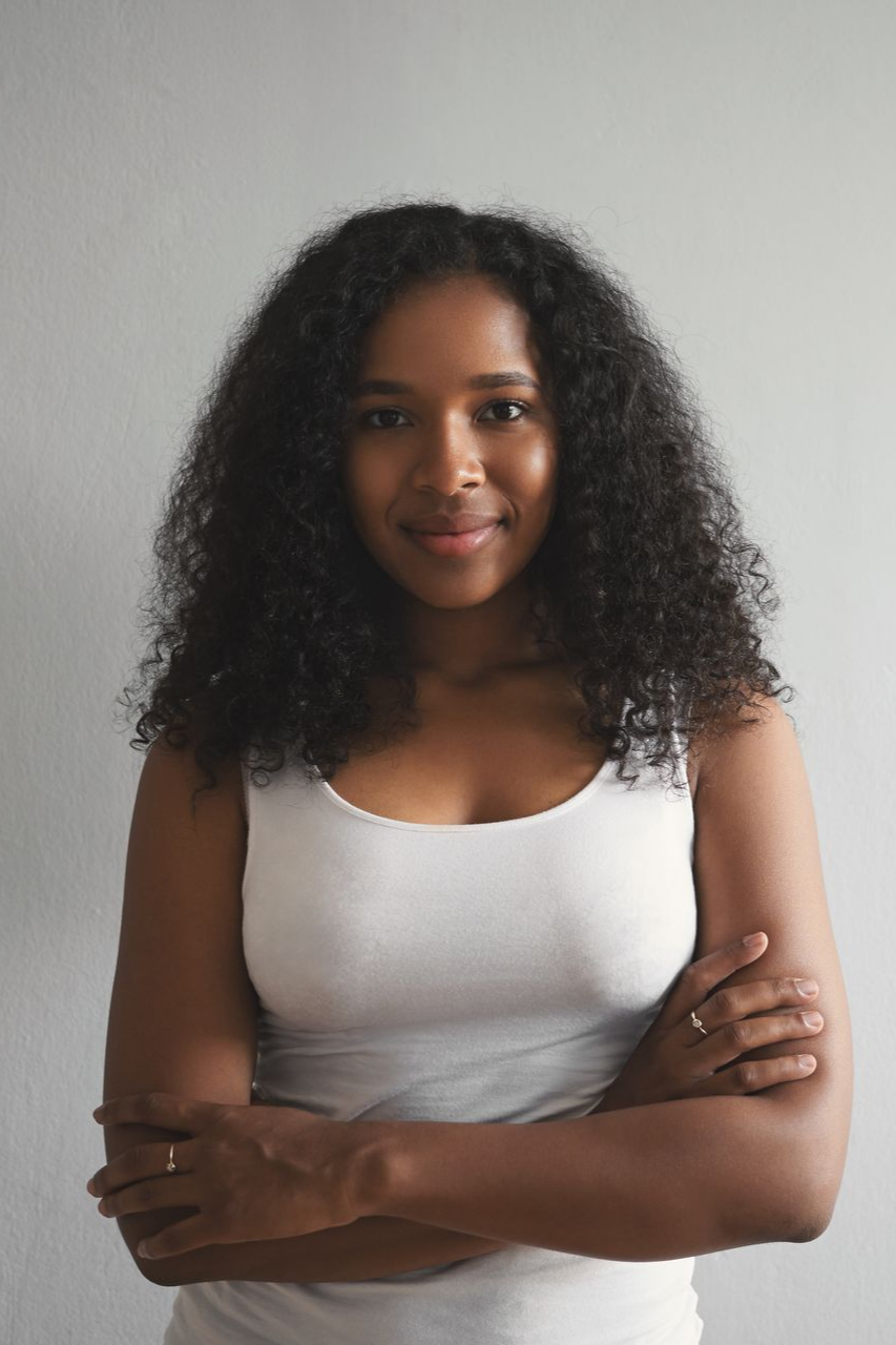 Woman with curly dark hair and arms crossed, wearing a white tank top, smiling against a plain grey background.