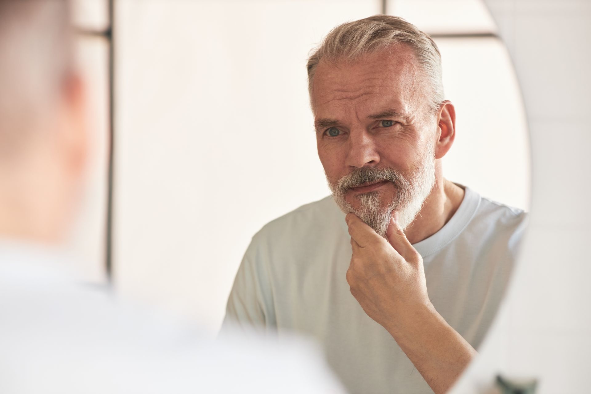 Man with gray hair and beard looking in a mirror, touching his chin in a bright bathroom