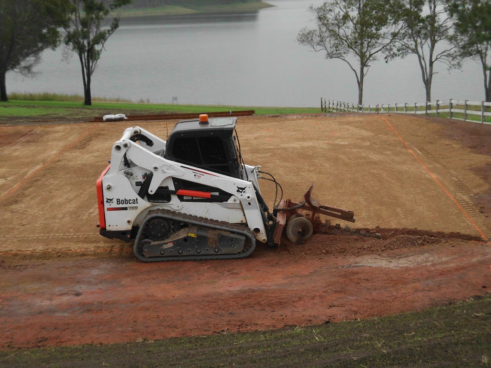 Bobcat Skid-Steer with A Tiller Attachment on Red Soil, Near a Lake with Trees and White Fence — Evans Earthmoving Contractors In Malanda, QLD