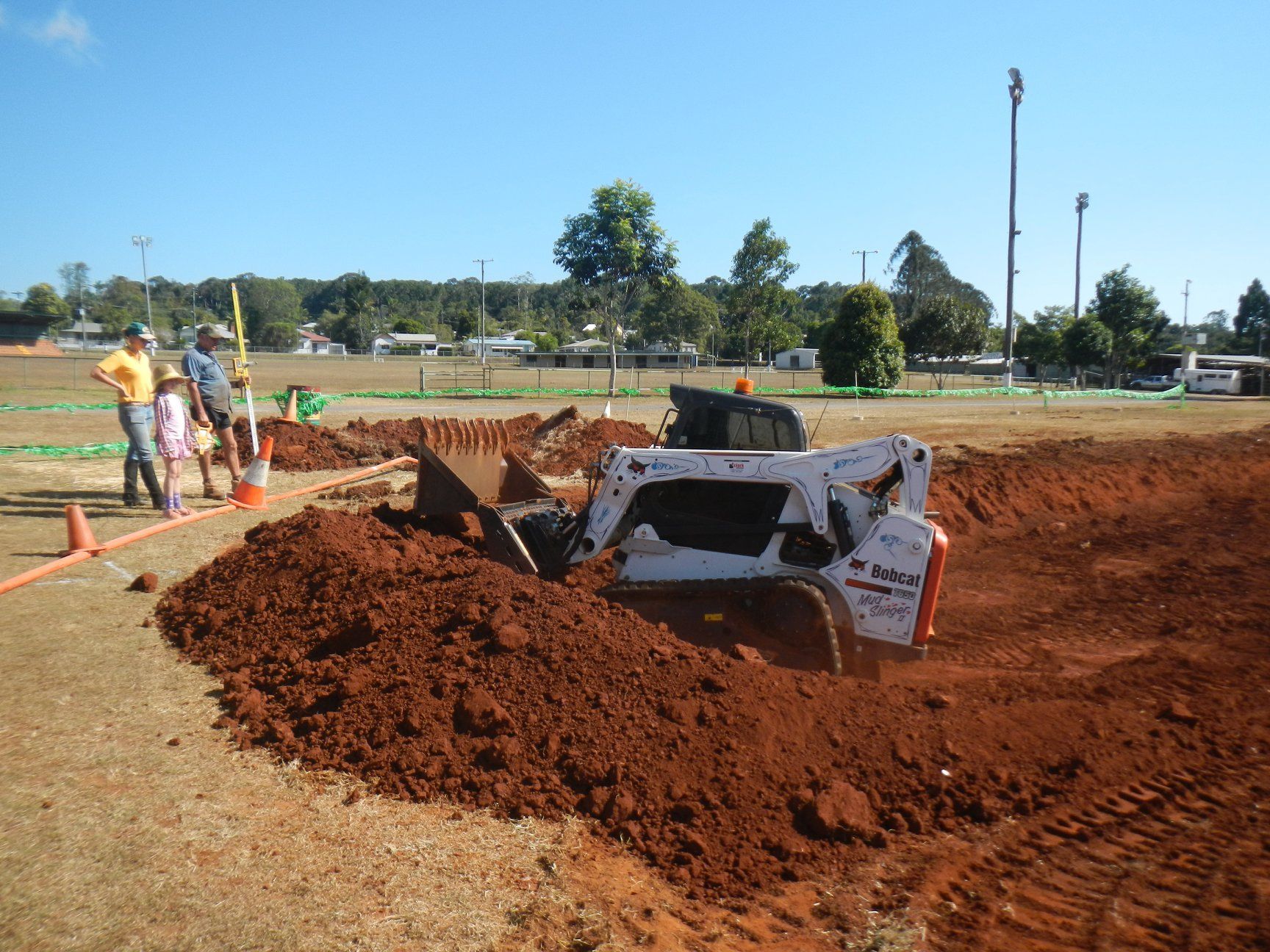 A Bobcat Compact Loader Digging in Red Soil on A Field with People Watching Under a Clear Sky — Evans Earthmoving Contractors In Malanda, QLD 