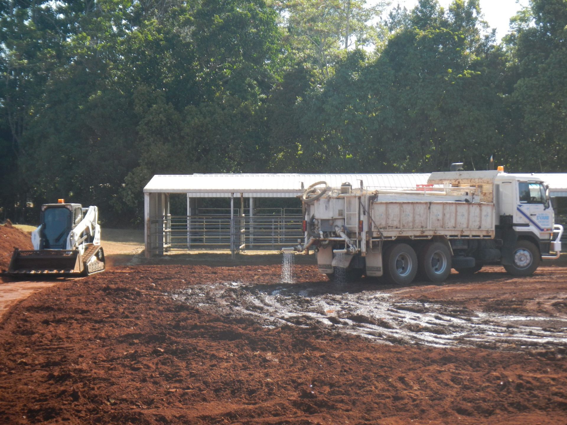 A Truck Dumping Material onto Muddy Ground with A Small Tractor Nearby, in Front of A Shed — Evans Earthmoving Contractors In Malanda, QLD
