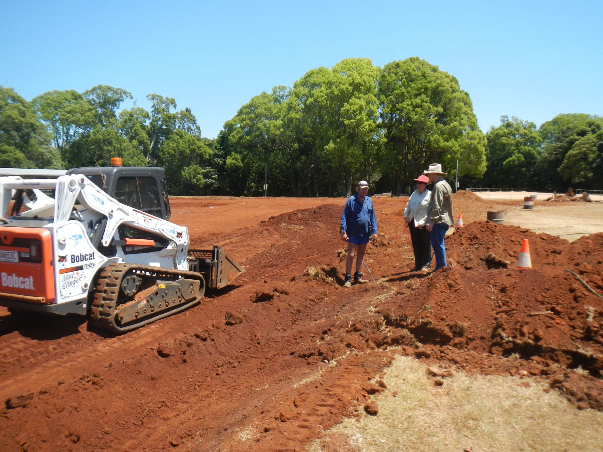 Bobcat Excavator Working on Red Dirt, Three People Observing, Sunny Day — Evans Earthmoving Contractors In Malanda, QLD