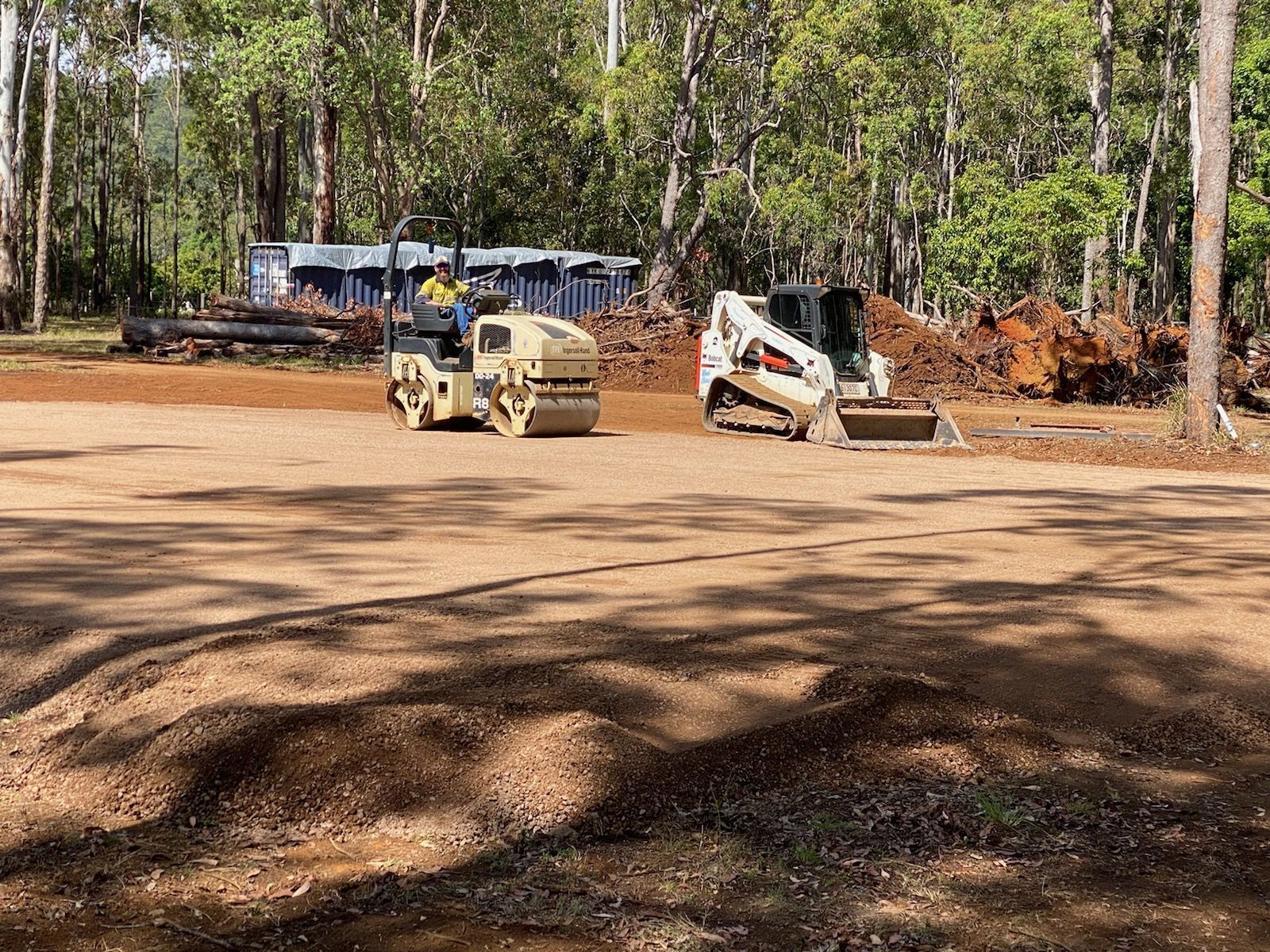 A Road Roller Compacts Gravel Next to A Skid-Steer Loader at A Construction Site in A Wooded Area — Evans Earthmoving Contractors In Malanda, QLD 