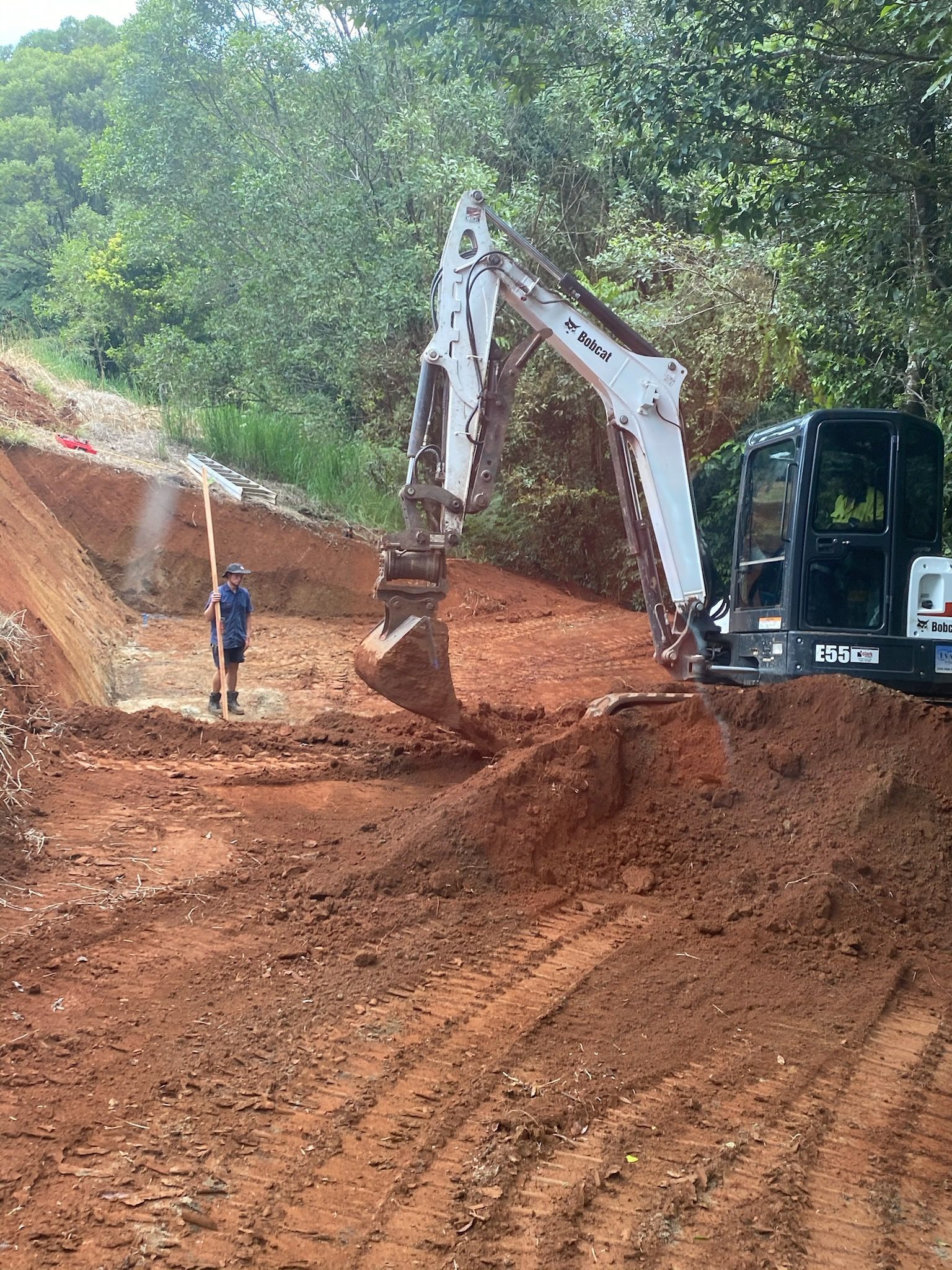 Excavator Digging Red Soil with A Person Observing in A Wooded Area — Evans Earthmoving Contractors In Malanda, QLD 