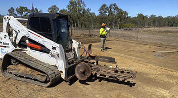 White Bobcat Trencher Digging a Ditch, with A Worker Observing on A Dusty Field Under a Clear Sky — Evans Earthmoving Contractors In Malanda, QLD
