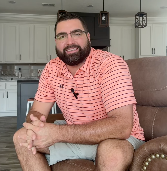 Man with beard in pink striped shirt sits on brown leather recliner in a kitchen, smiling.