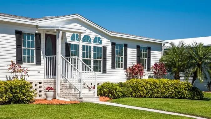 White manufactured home with black shutters, front porch, and landscaped yard.